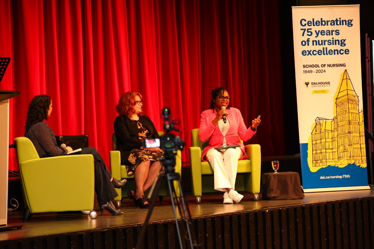 Three women seated in green chairs on stage with a backdrop of red curtains at an event while one speaks into a microphone.