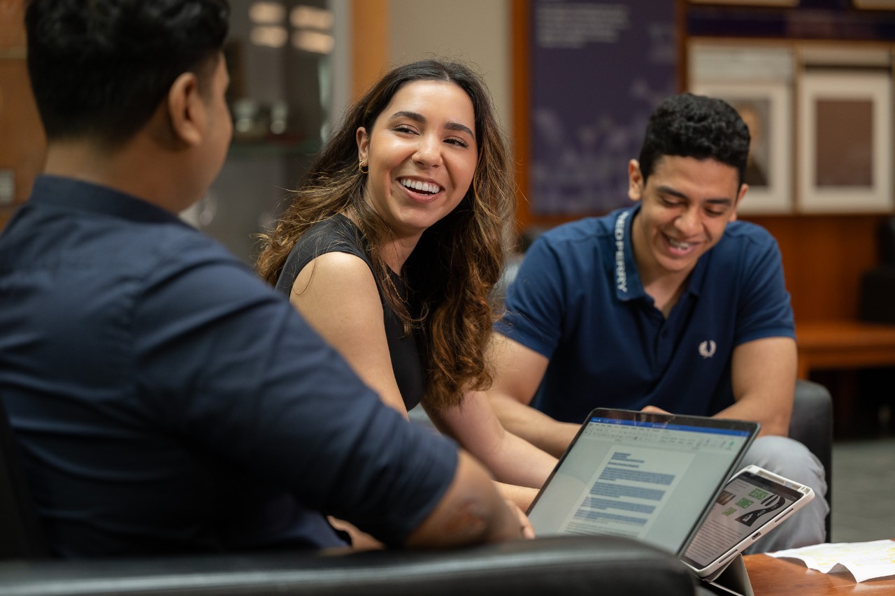 A female student sits with two others, smiling and working on laptops on campus.