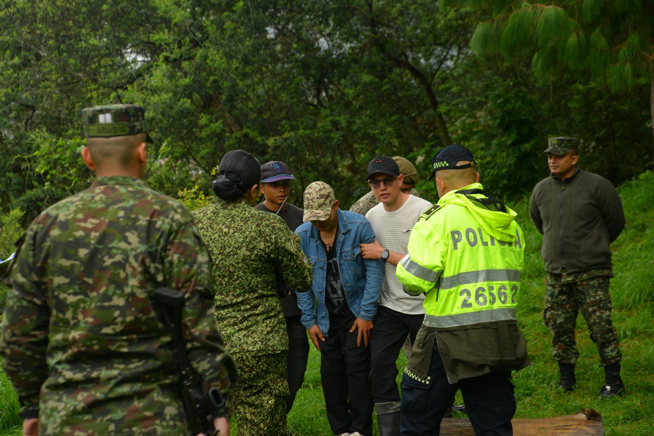 Police and military personnel depict detaining a group of people in an outdoor, wooded area.