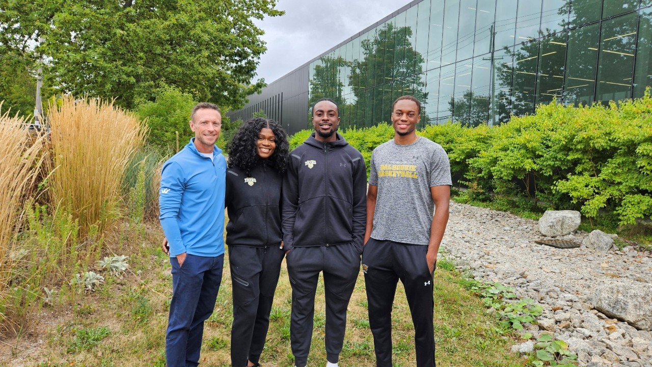 A man stands with three university students, all in athletic wear, alongside a path with greenery outside a building with a wall of windows.