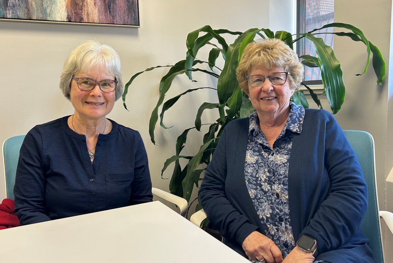 Two women seated at a table smiling in a small bright office space with a large plant.