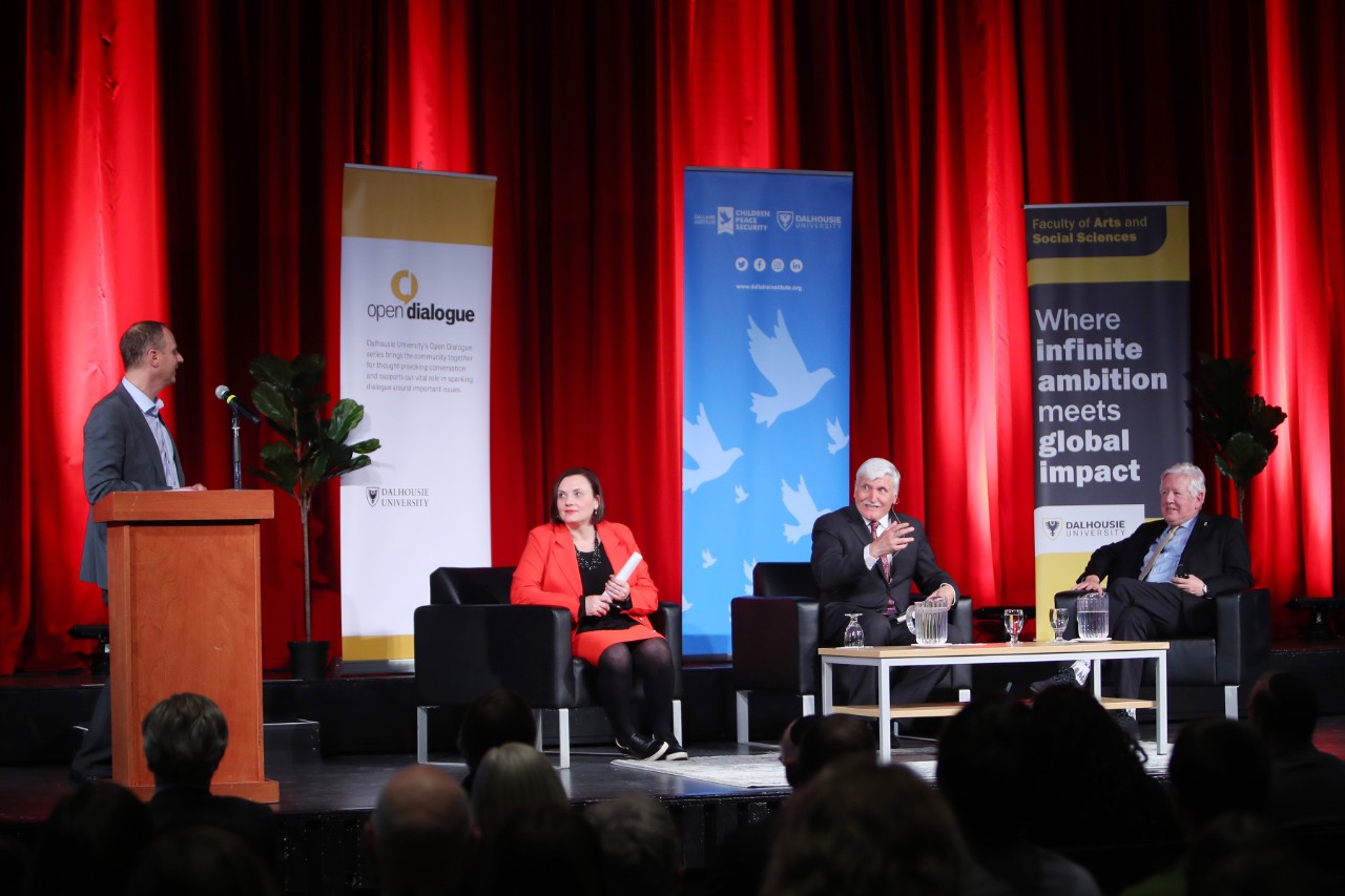 A man at a podium speaks to three people seated in chairs on a stage