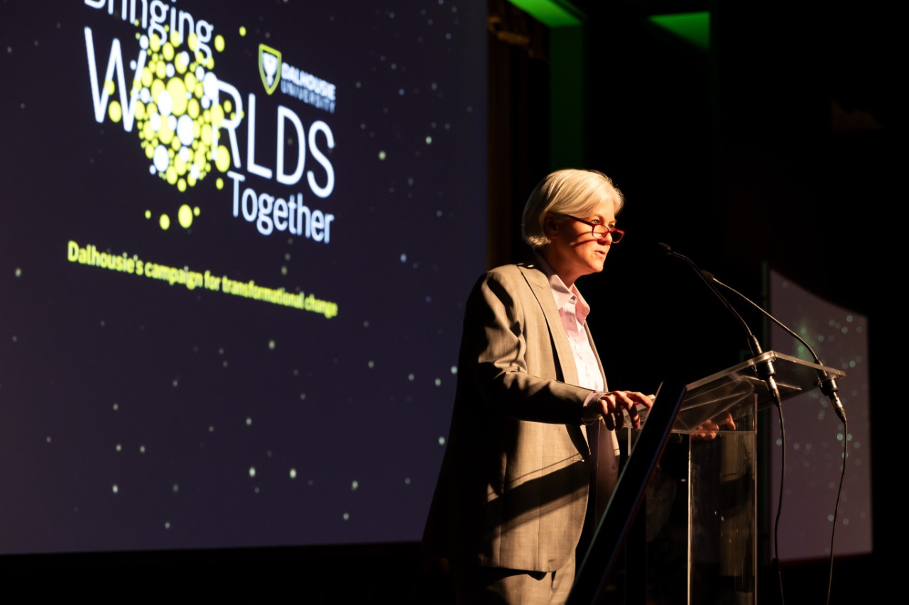 Dalhousie President Kim Brooks stands at a podium, looking out into the crowd. The Bringing Worlds Together logo is displayed on a screen behind her.