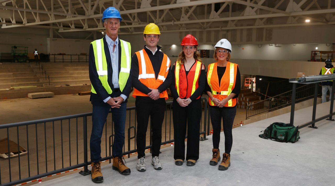 Two donors stand with two students smiling together wearing hard hats, boots and reflective vests in a building under construction.