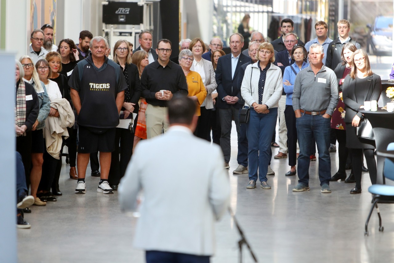 A man stands talking to a crowd gathered around in an open, light interior space.