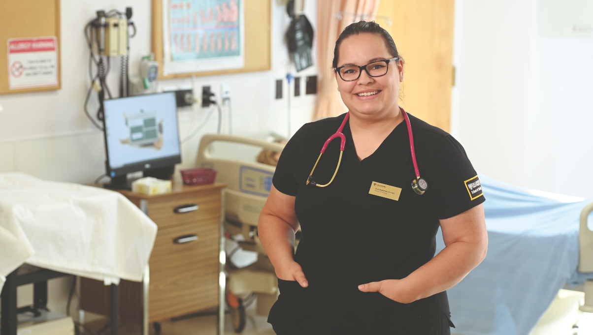 A woman wearing glasses and black scrubs with a pink stethoscope standing in a hospital room