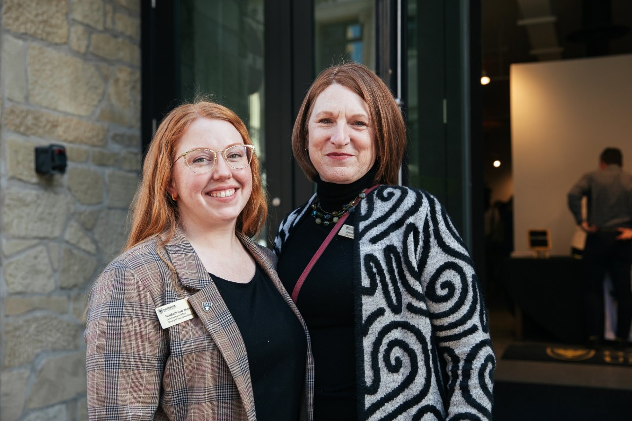 Two women stand smiling together outside the entrance to an event venue.