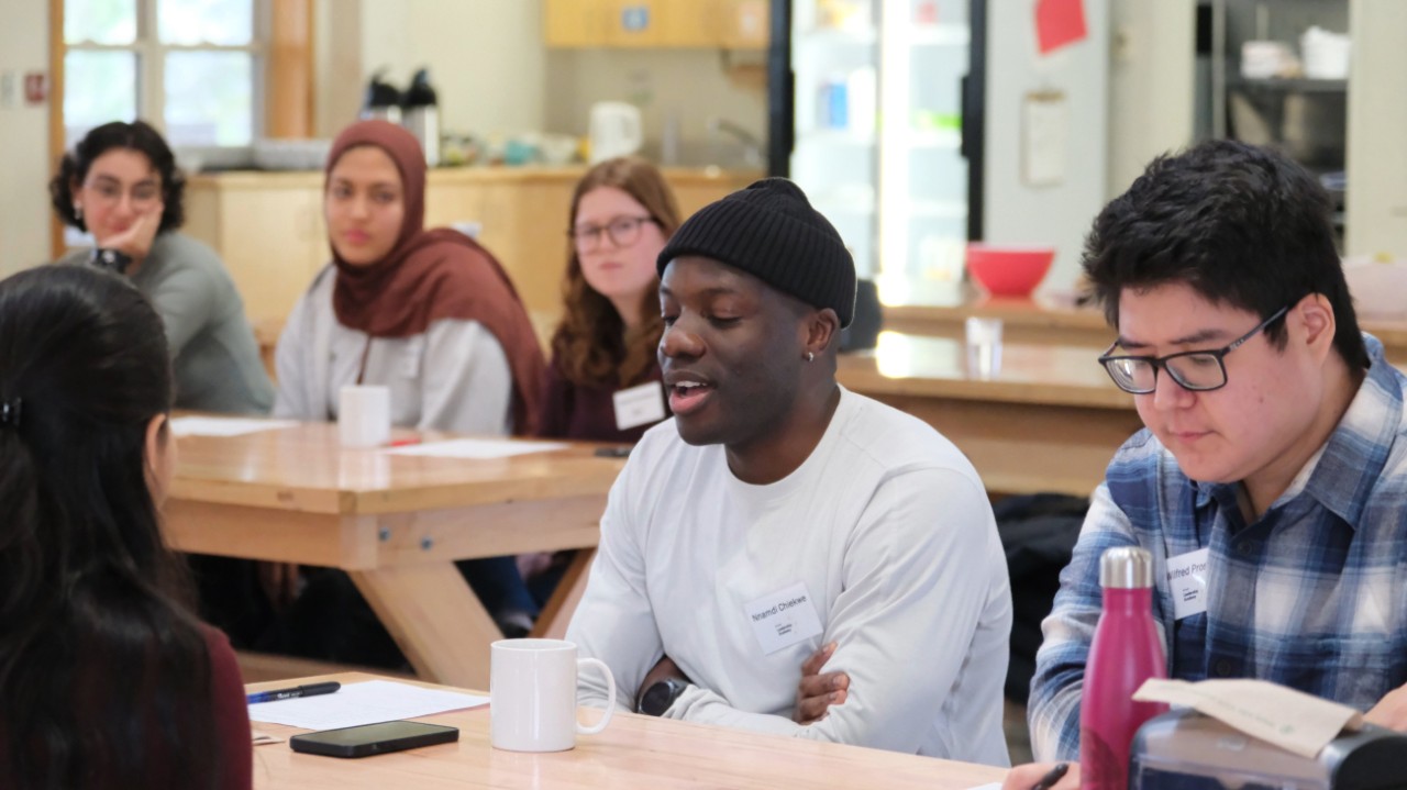 A diverse group of students seated at wooden tables in a casual indoor setting.