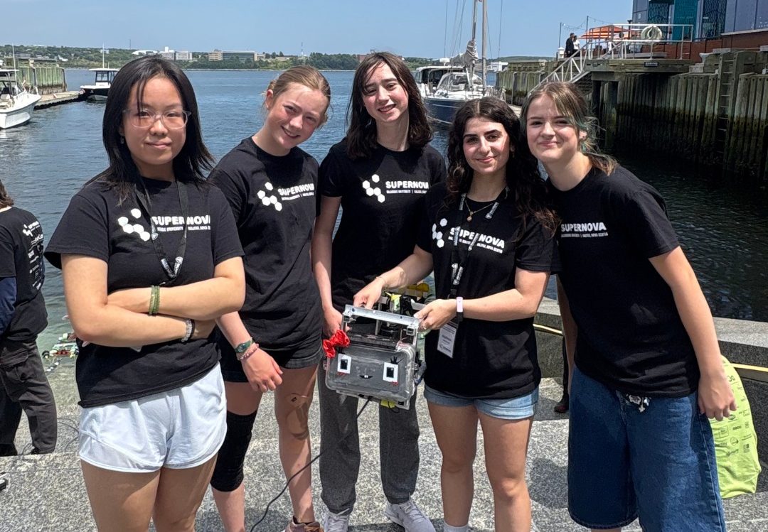 A group of five students wearing black t-shirts on the city waterfront pier in fine weather holding a small mechanical device.