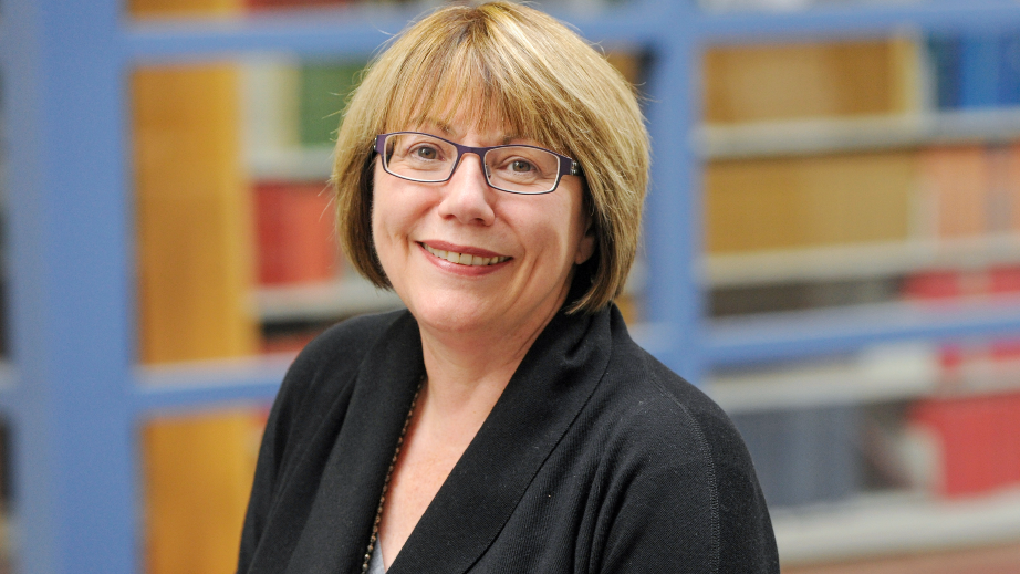 A woman with short blonde hair wearing glasses and a black blazer standing in a library
