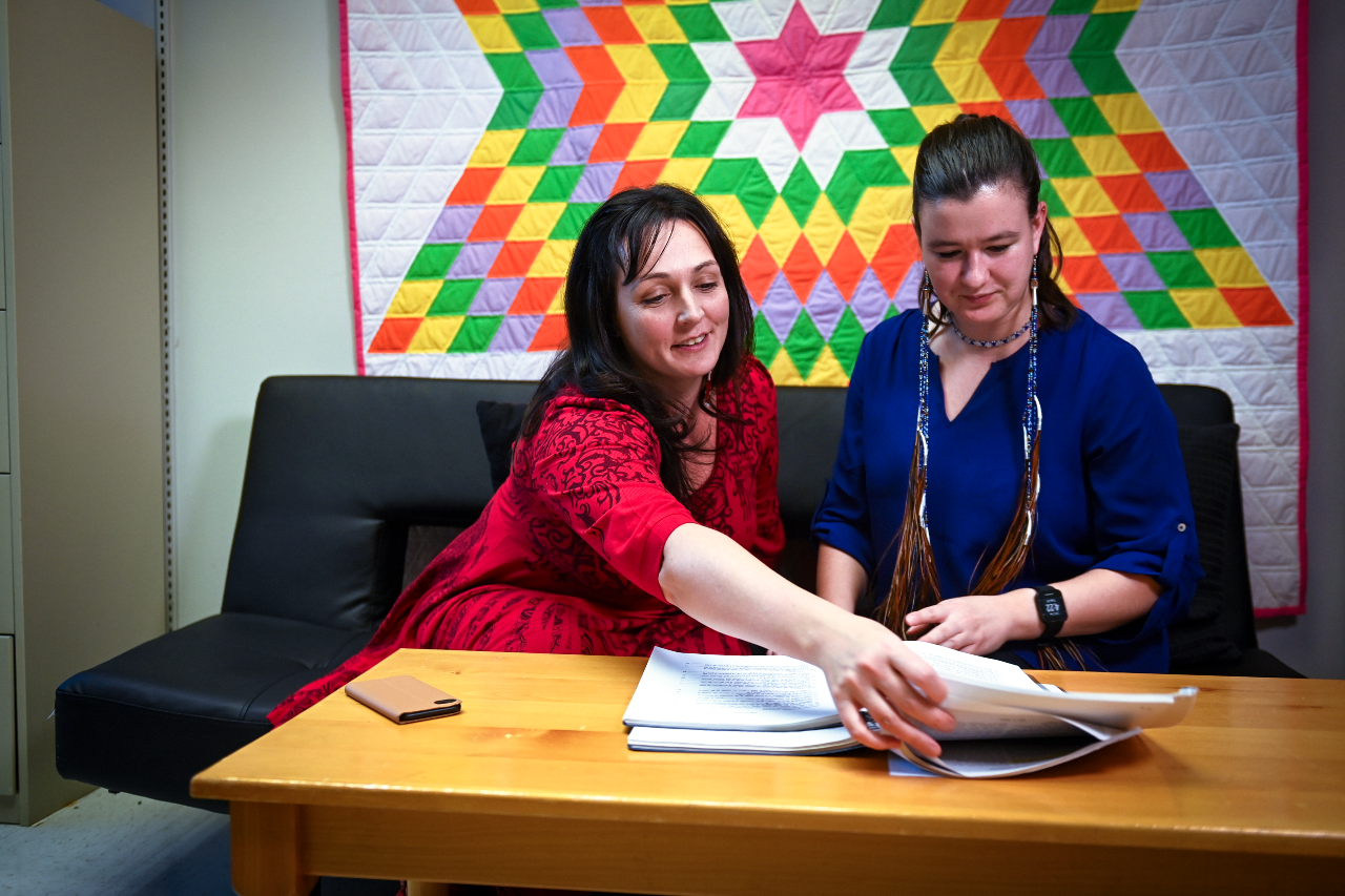 A woman wearing a red dress seated on a couch showing paperwork to a woman in a blue top 