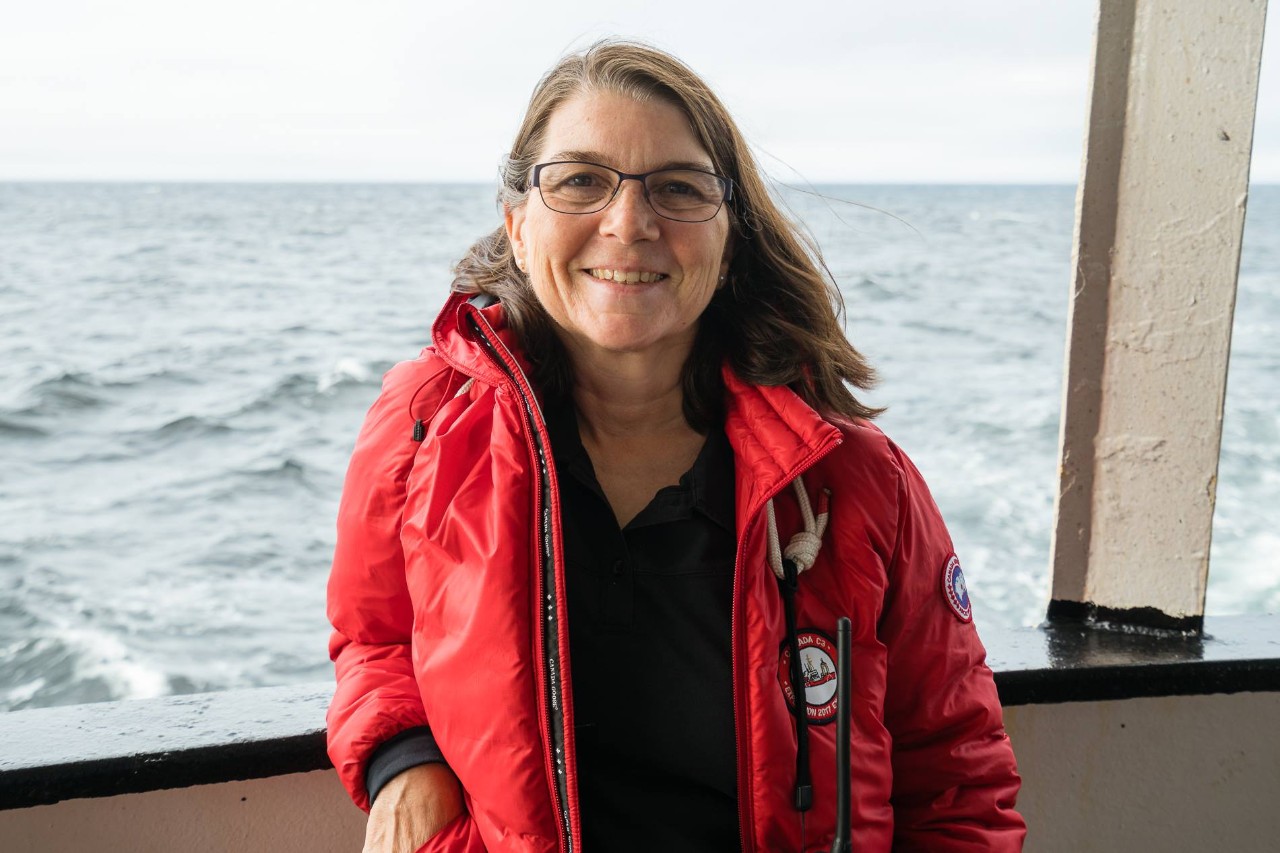 A woman wearing a red coat and glasses stands on the deck of a boat