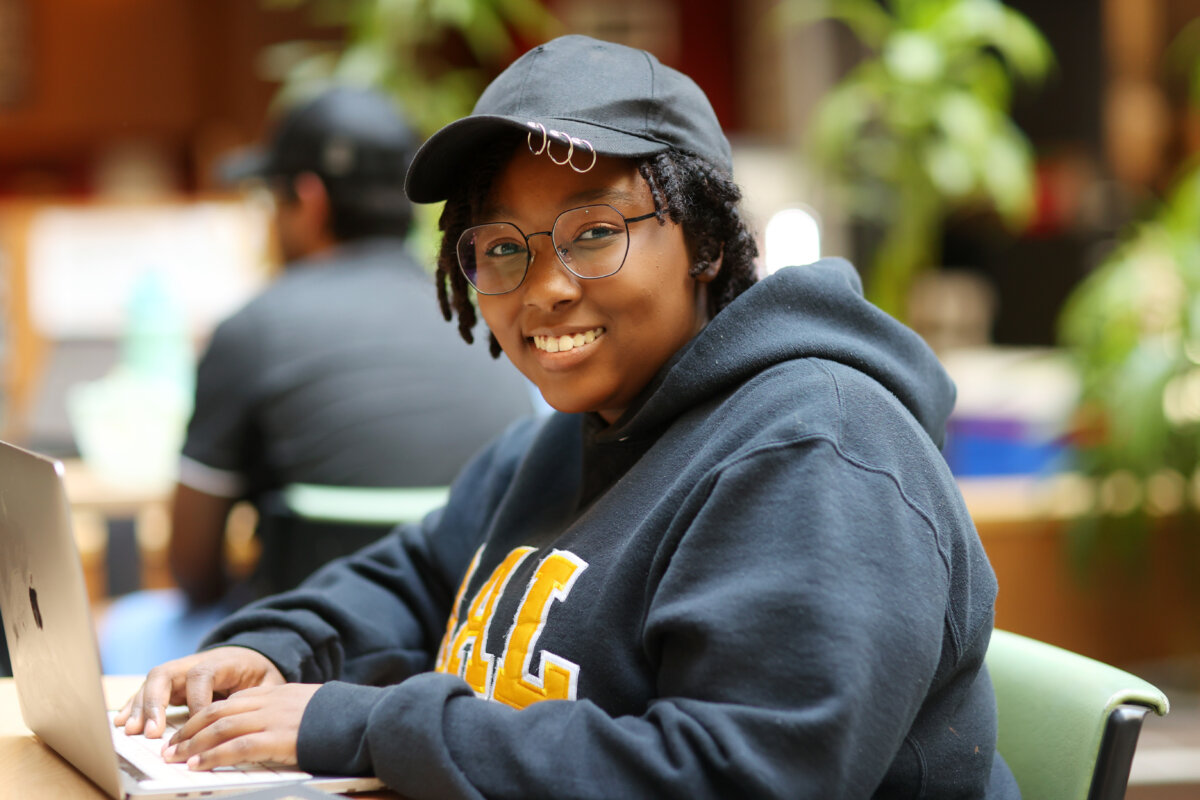 A university student wearing a black baseball cap and DAL hoodie sits smiling with hands poised above a MacBook. 