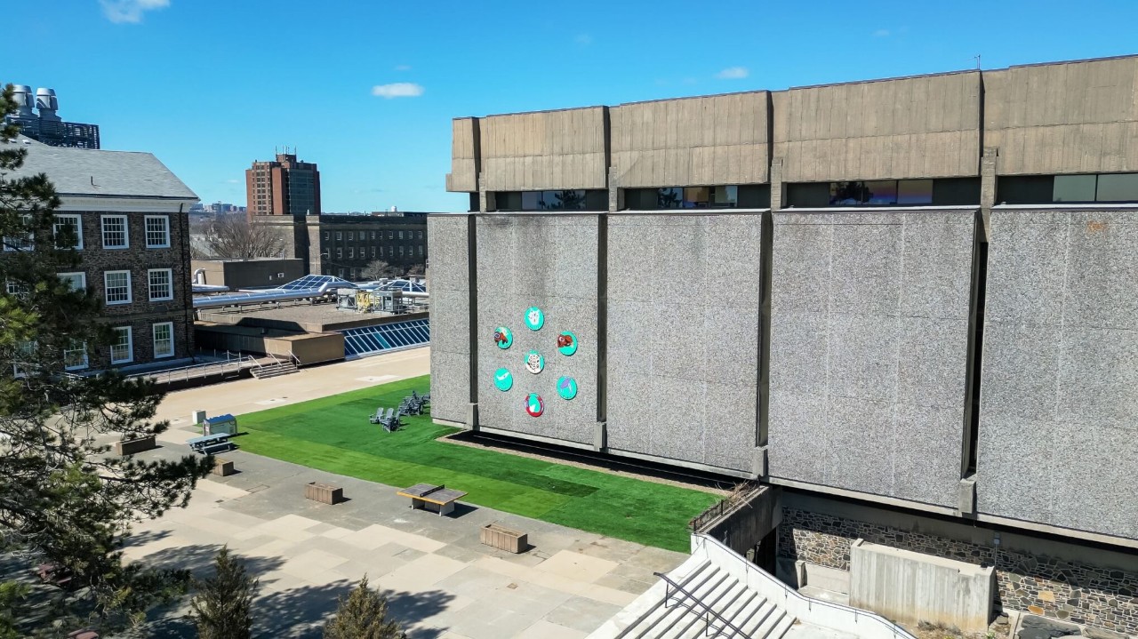 A modern building features colorful artwork on its concrete wall, surrounded by green grass and outdoor seating in a bright, sunny courtyard.
