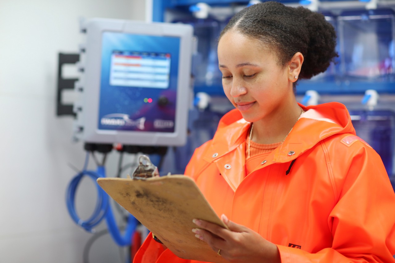 A female-presenting person is wearing an orange raincoat and holding a clipboard in a lab with a monitor in the background.