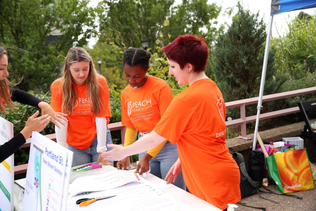 Four students wearing bright orange "PEACH Research Unit" t-shirts collaborate at an outdoor table covered with materials with a backdrop of greenery.