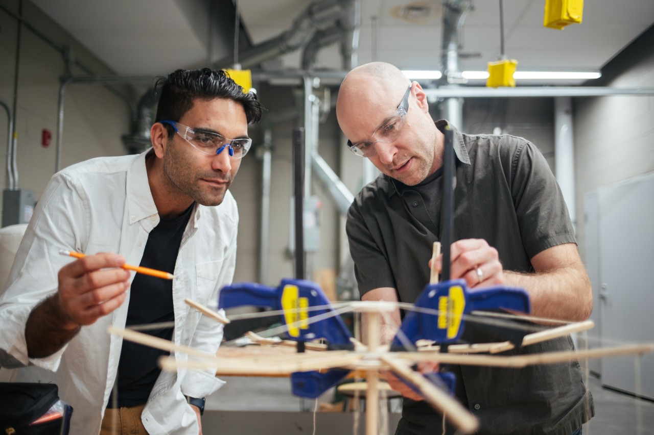 Two people examine a small wooden structure that resembles an umbrella