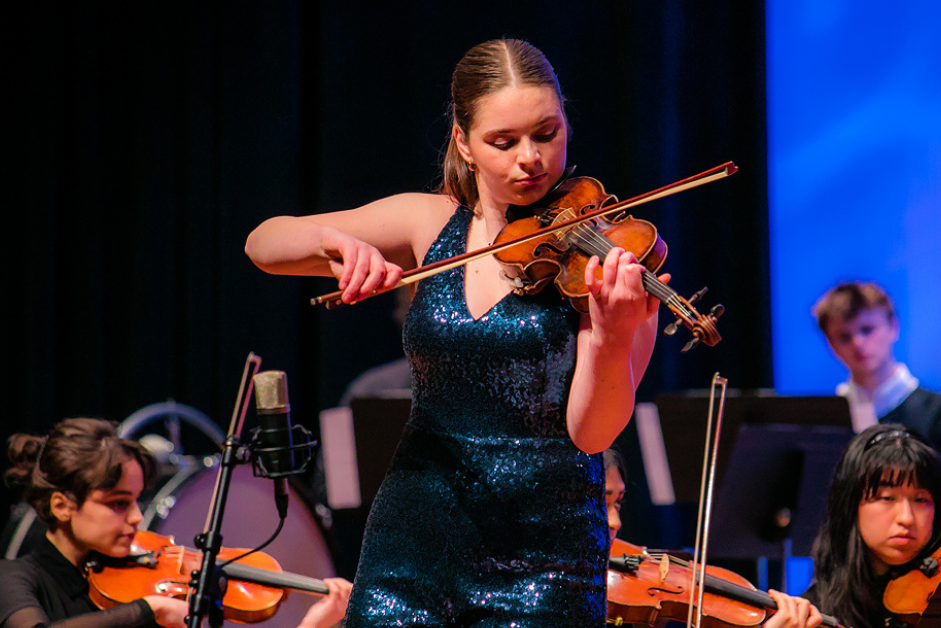 A woman wearing a blue sequined dress plays a violin on stage with other violinists