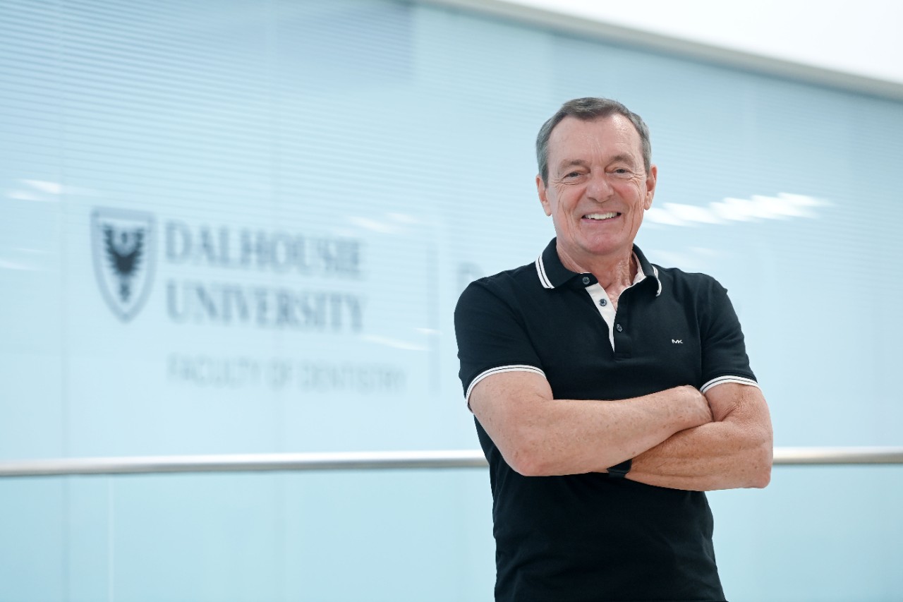 Boran in a black short-sleeved collared shirt standing with his arms crossed smiling in the hallway of the dentistry building.