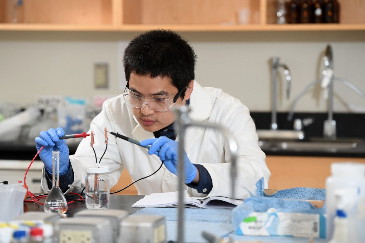 A man wearing a white lab coat, safety glasses and blue gloves works on a scientific engineering experiment.