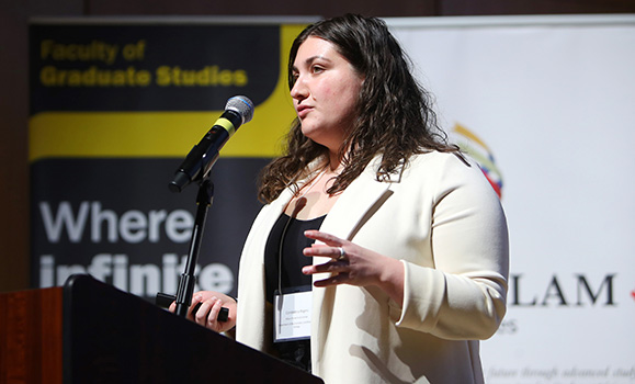 Rogers wearing a cream-coloured blazer speaking into a mic at a podium during the event with Dalhousie and Killam banners in the background.