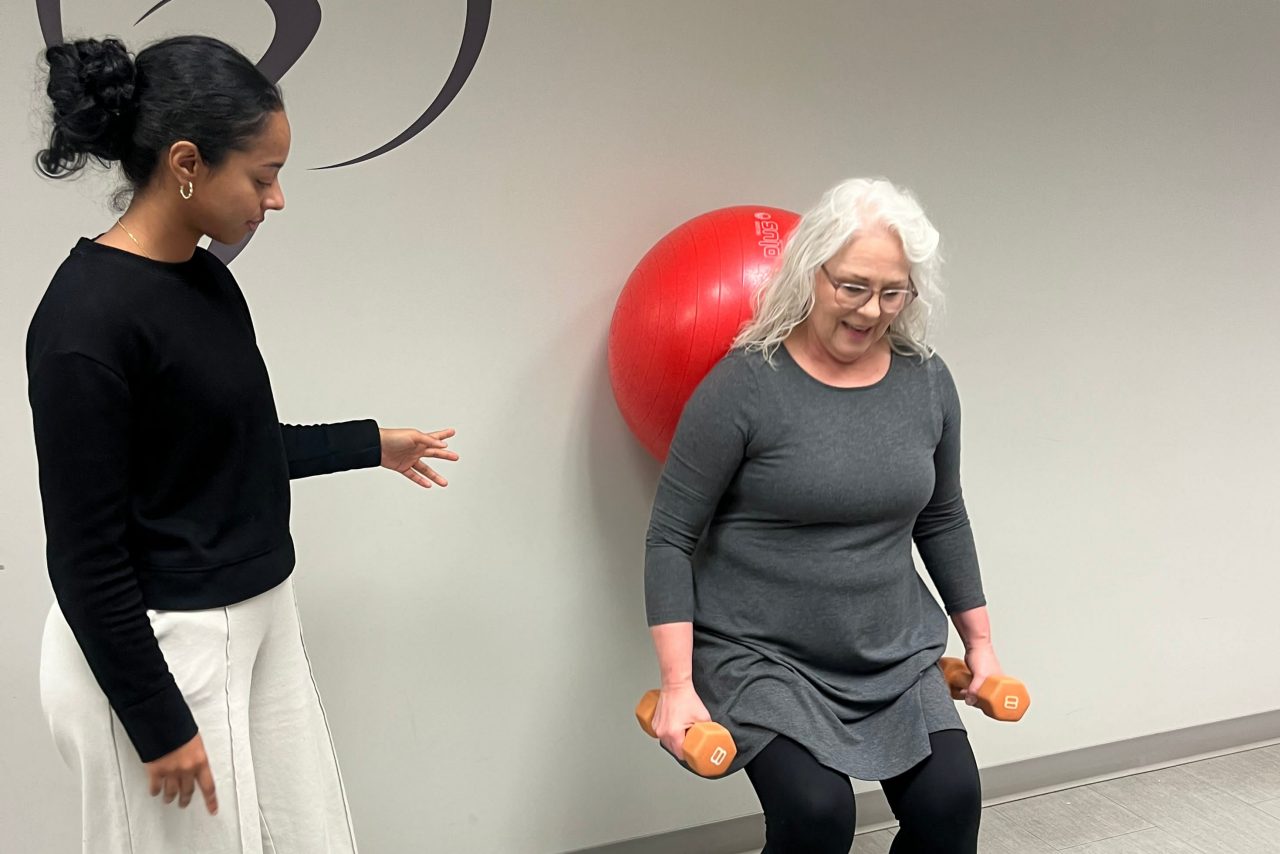 Cherian assists a patient in the clinic who is using a balance ball on the wall with hand weights.