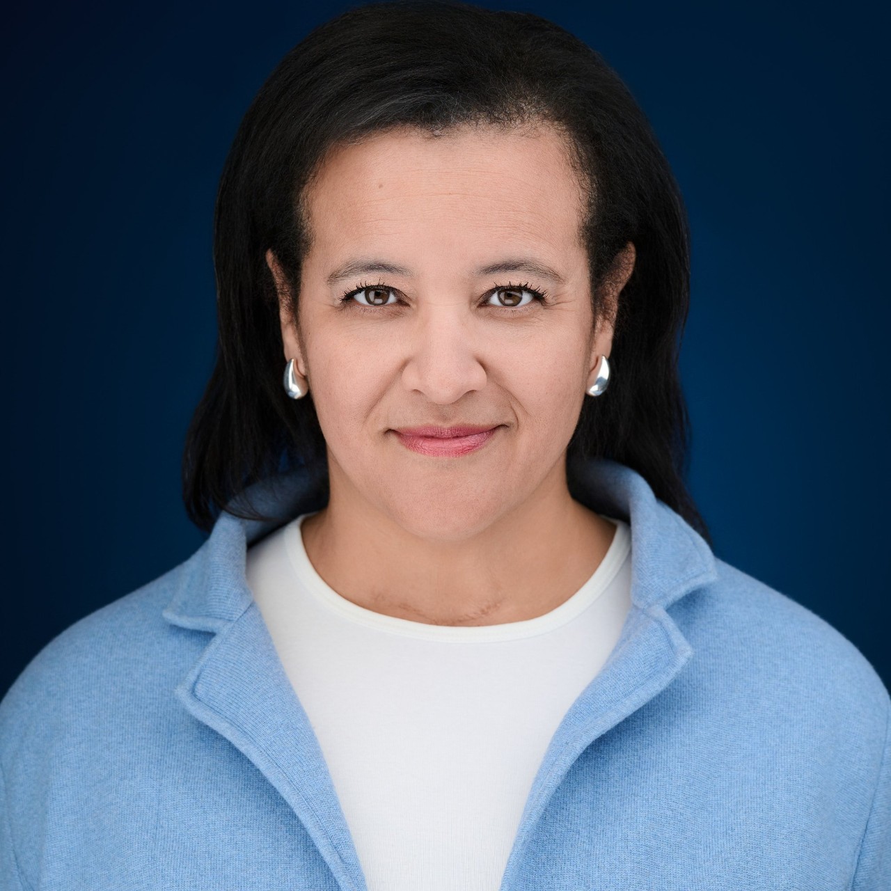 A woman wearing a white t-shirt and blue sweater looks at the camera