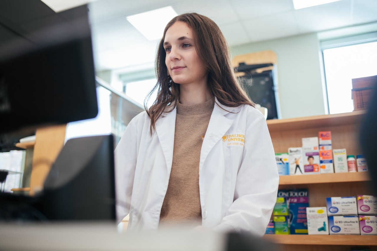 A woman wearing a white lab coat stands at a computer with pill boxes visible in the background