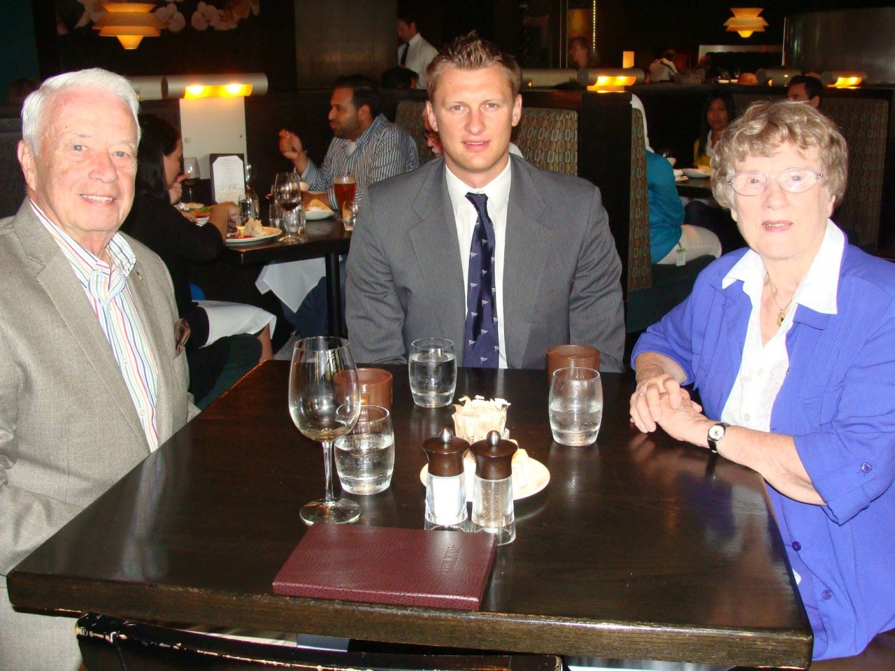 A younger man sits with a man and a woman, all dressed business casually, at a table with glasses on it in a restaurant.