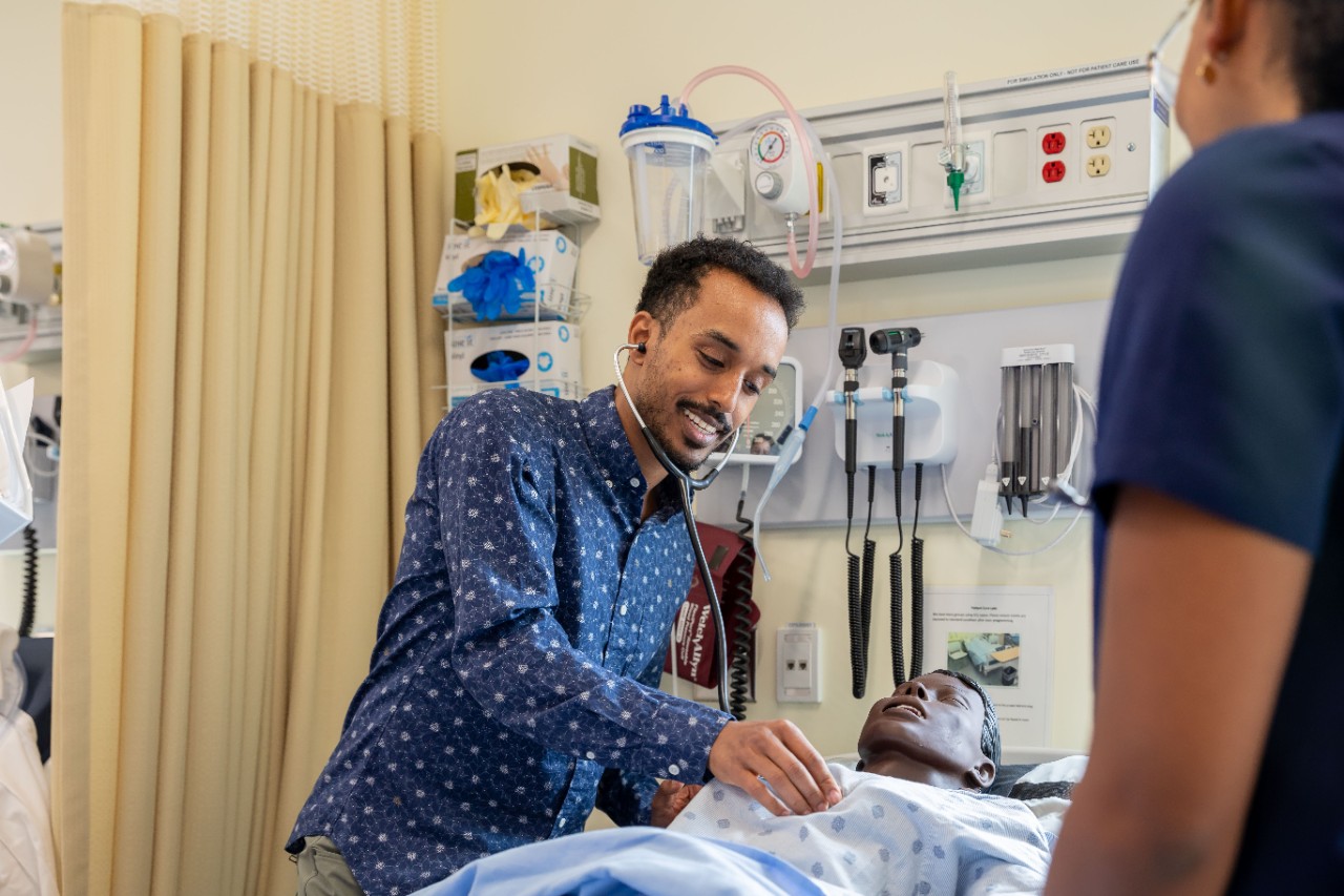 Student holds stethescope to chest of simulation mannikin as another student looks on.