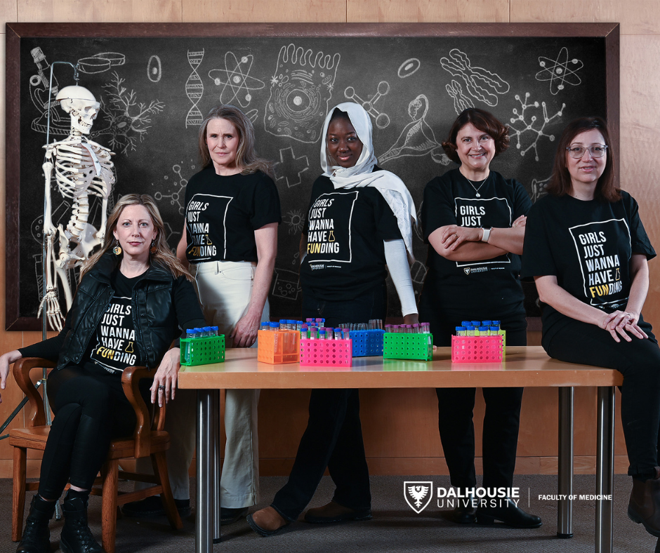 Five women in matching black t-shirts pose in a science classroom, surrounded by test tubes, a skeleton model, and a blackboard filled with science doodles. Three stand behind a desk, one sits in a chair, and another sits on the desk.