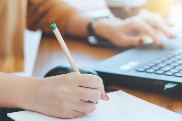 A person writes on a piece of paper atop a table while scrolling on a laptop with only their hands, arms and part of torso in view.