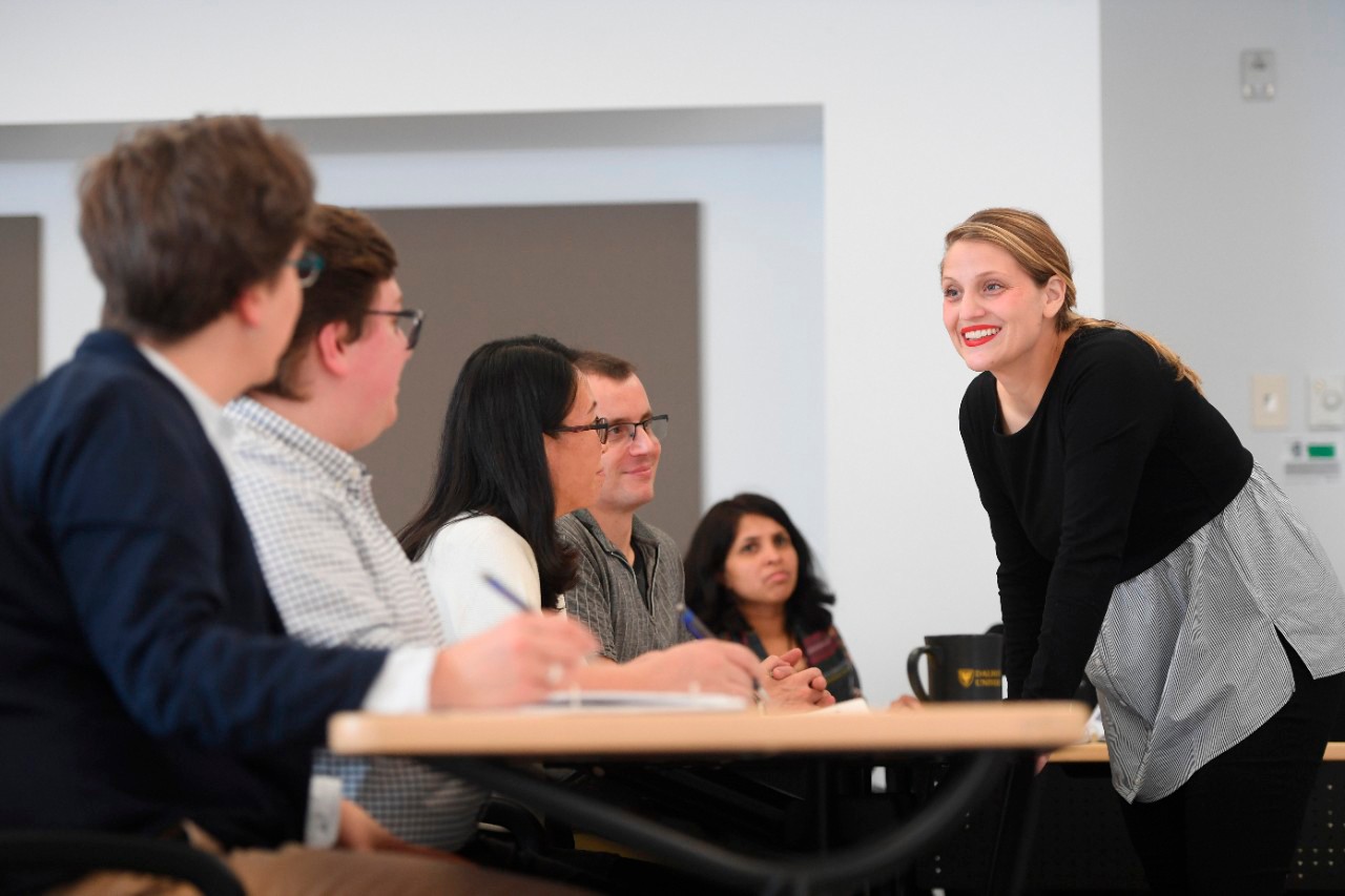 A diverse group of several students sitting at a classroom table looking towards a woman instructor leaning on the table and smiling back at them.