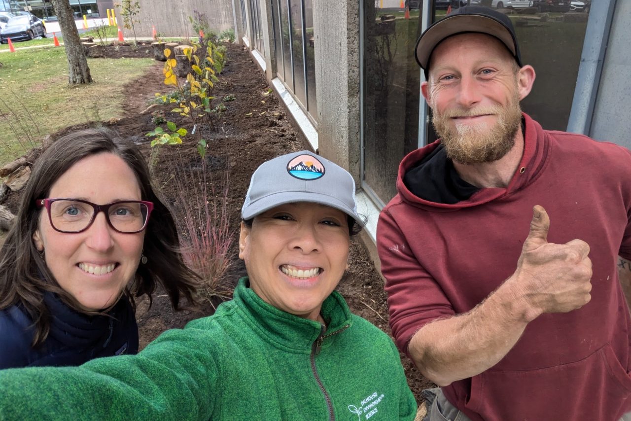 A selfie of three people with one giving a thumbs up in front of a garden outside of a campus building.