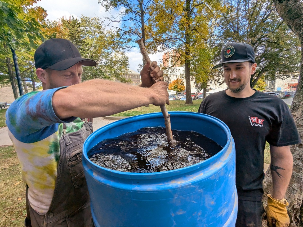 A man stirs a large container of water and soil while another looks on outdoors in fine weather.