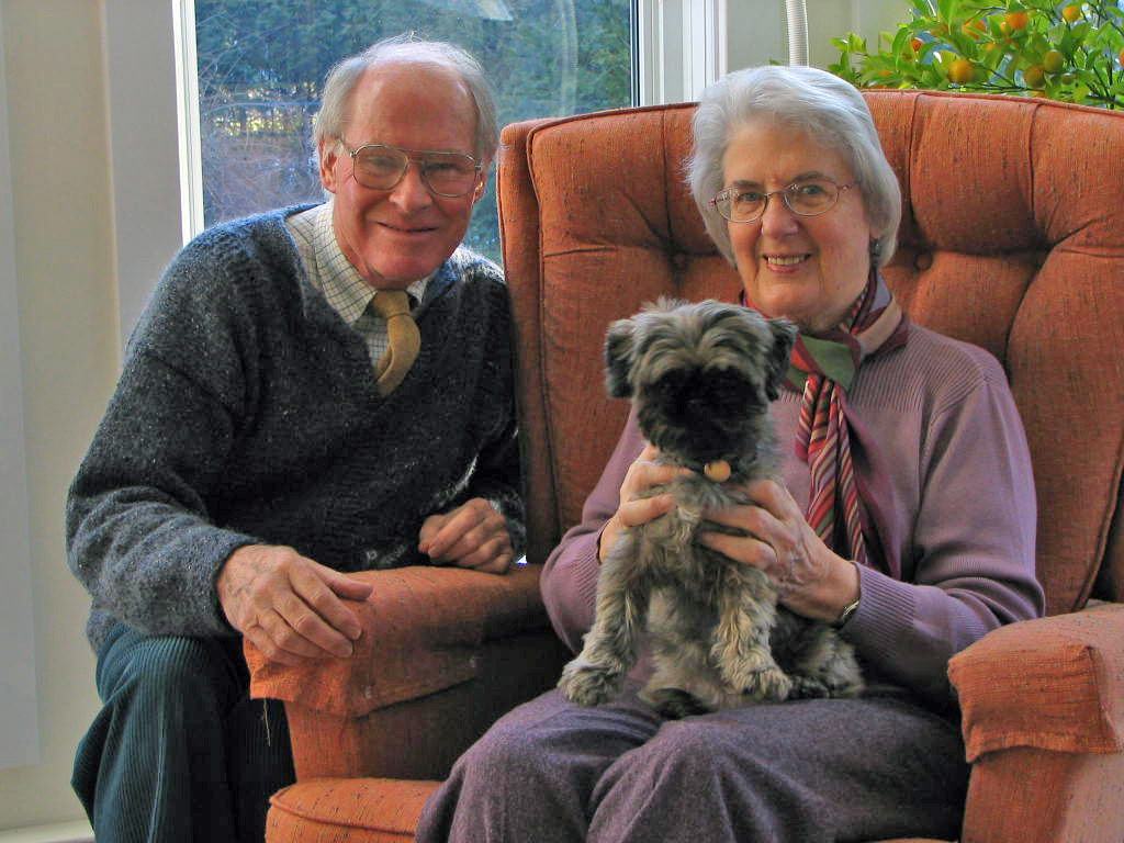 A woman is seated in a comfortable chair holding a small dog on her lap while a man leans in beside her, smiling.