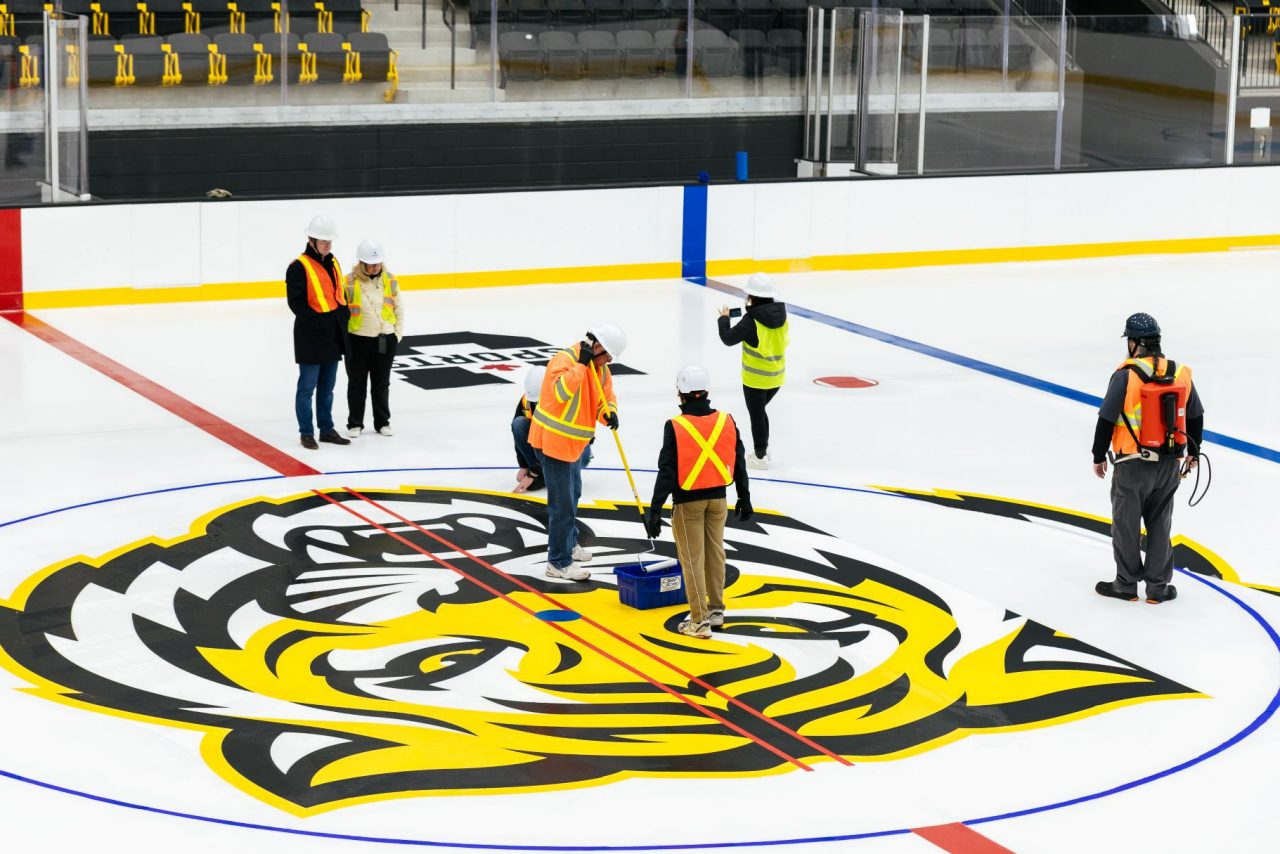 Workers in fluorescent vests and hard hats stand on large tiger logo in the middle of an indoor ice rink.
