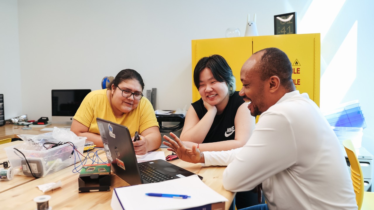 Three students collaborate on a laptop while seated around a table.
