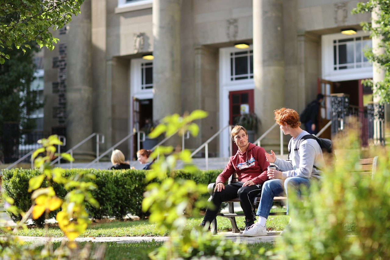 Two casually dressed male students sit on a bench with greenery in the foreground and a stone academic building with columns in the background.