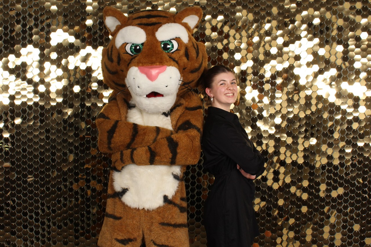A student stands in front of a sparkly gold background with her back leaning against a Tiger mascot. Both have their arms crossed.