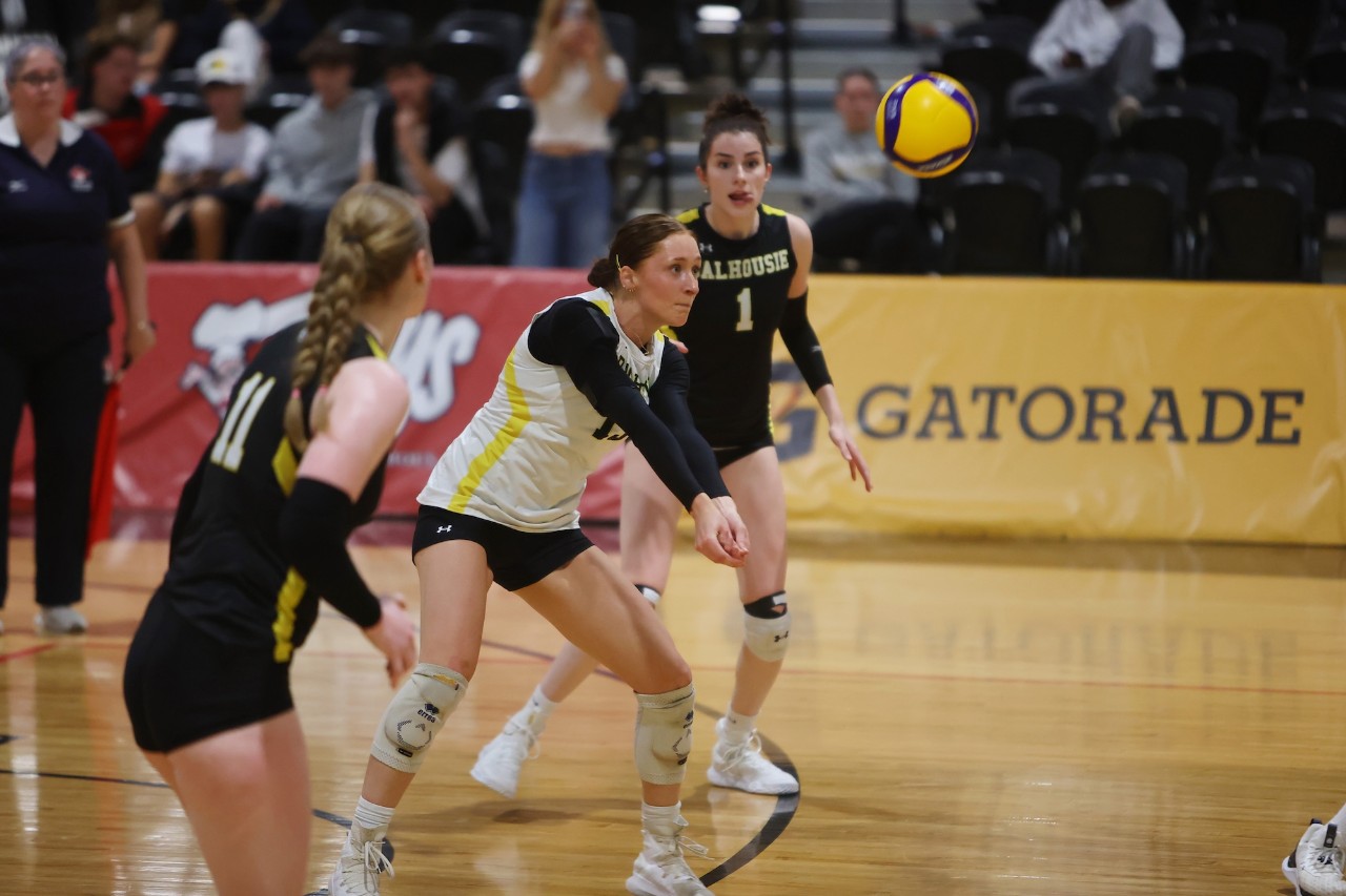 Three Dalhousie women's volleyball players are on the court as the one in the middle bumps the yellow volleyball into the air.