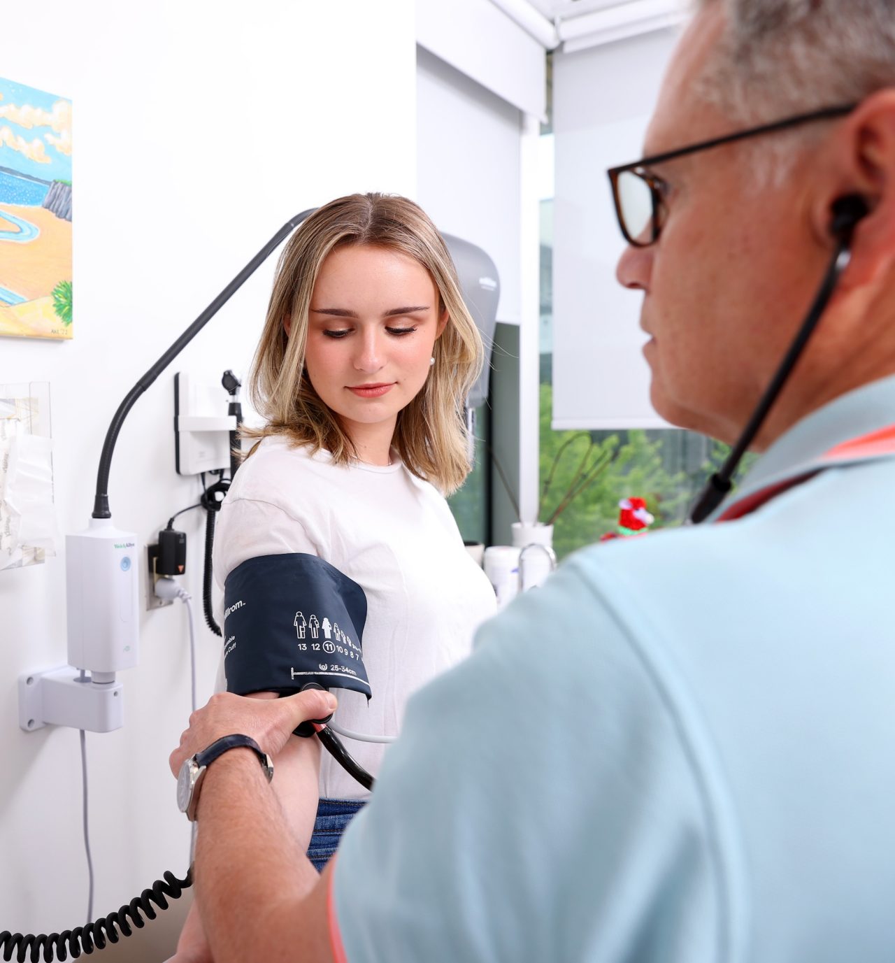 A student sits with a blood pressure cuff on her arm while a doctor wearing glasses and stethoscope holds her arm.