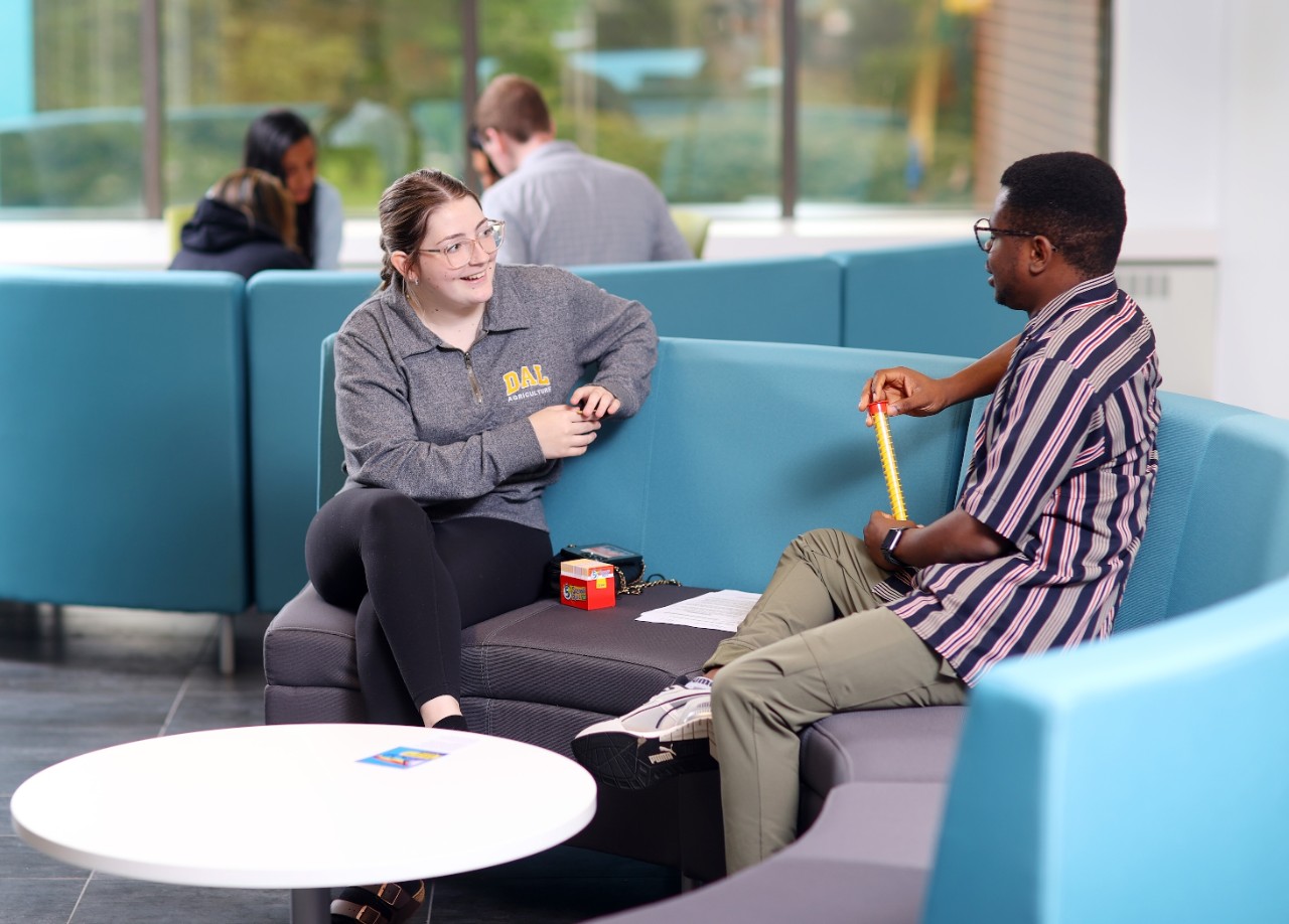 Two people sit and talk on a light blue, curved, padded bench with a small round table in front of them. In the background is a group of people sitting on a similar set of seats.