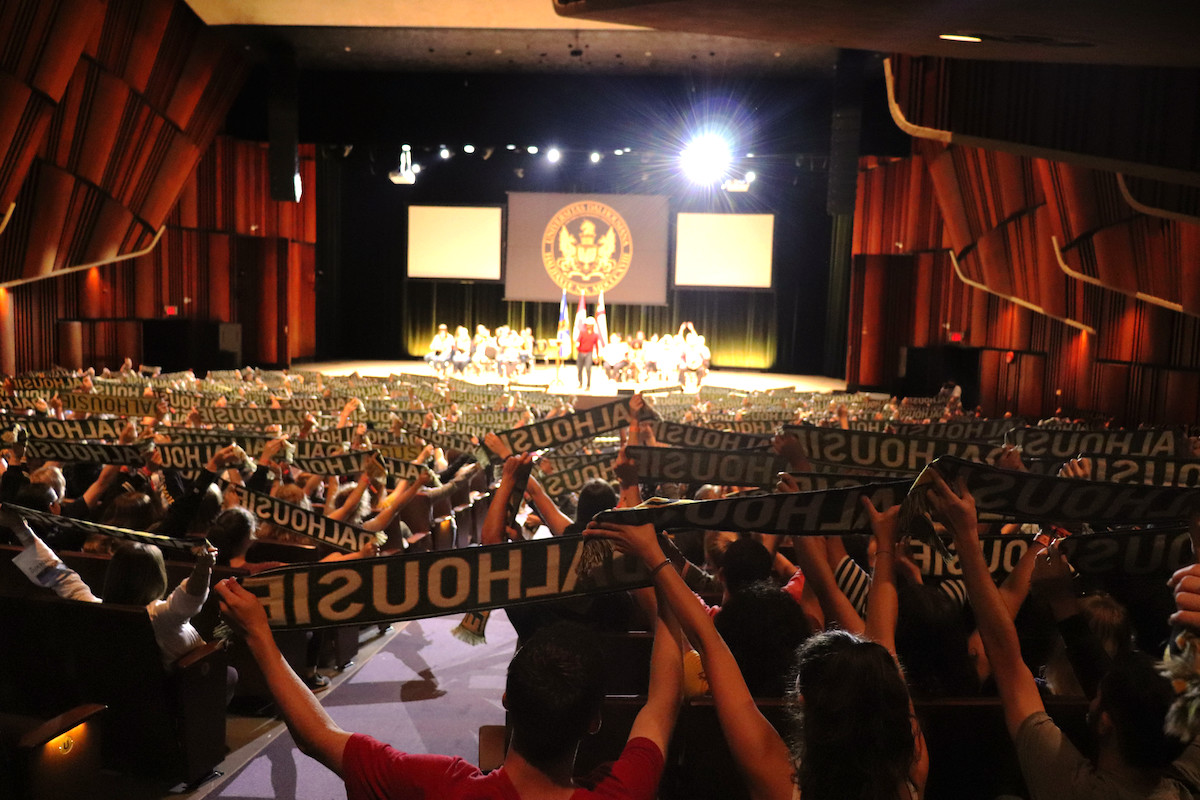 A theatre is filled with people in the seats holding up scarves that have Dalhousie written on them. There are people on stage in the distance.