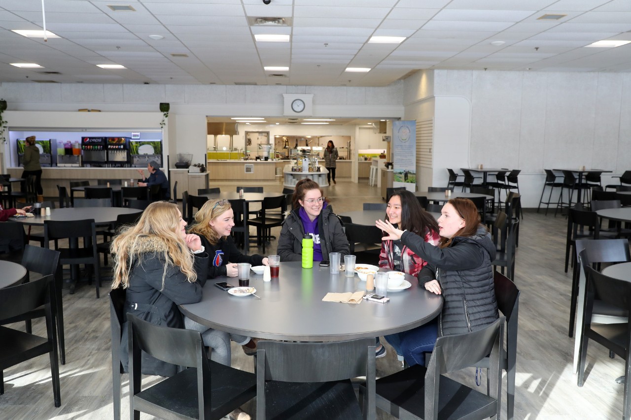 Five students sit around a circular table in Jenkins Hall on the Truro campus, eating a variety of foods for lunch, each with drinks.  