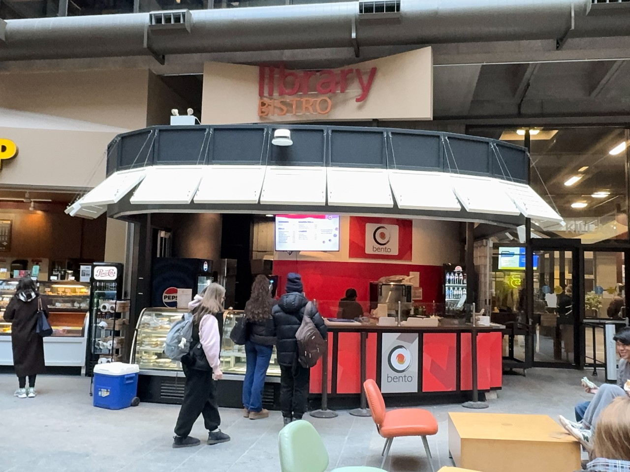 The Bento location in the Killam has a decorative roof with white shutters and a red rounded counter. There is a case of pre-prepared food on the left and a counter for fresh food on the right