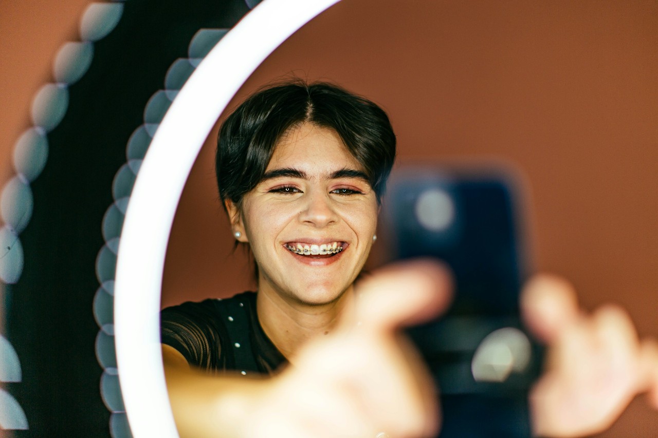 A young non-binary student with dark hair and braces takes a selfie using a ring light