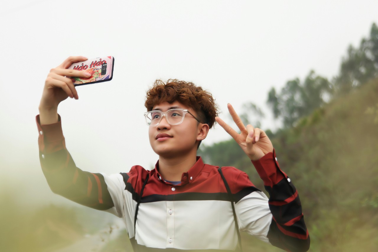 A young asian man with glasses and curly hair takes a selfie with a smartphone