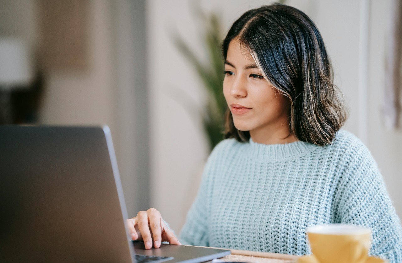 A young woman looks at her open laptop, with a coffee on the desk beside her