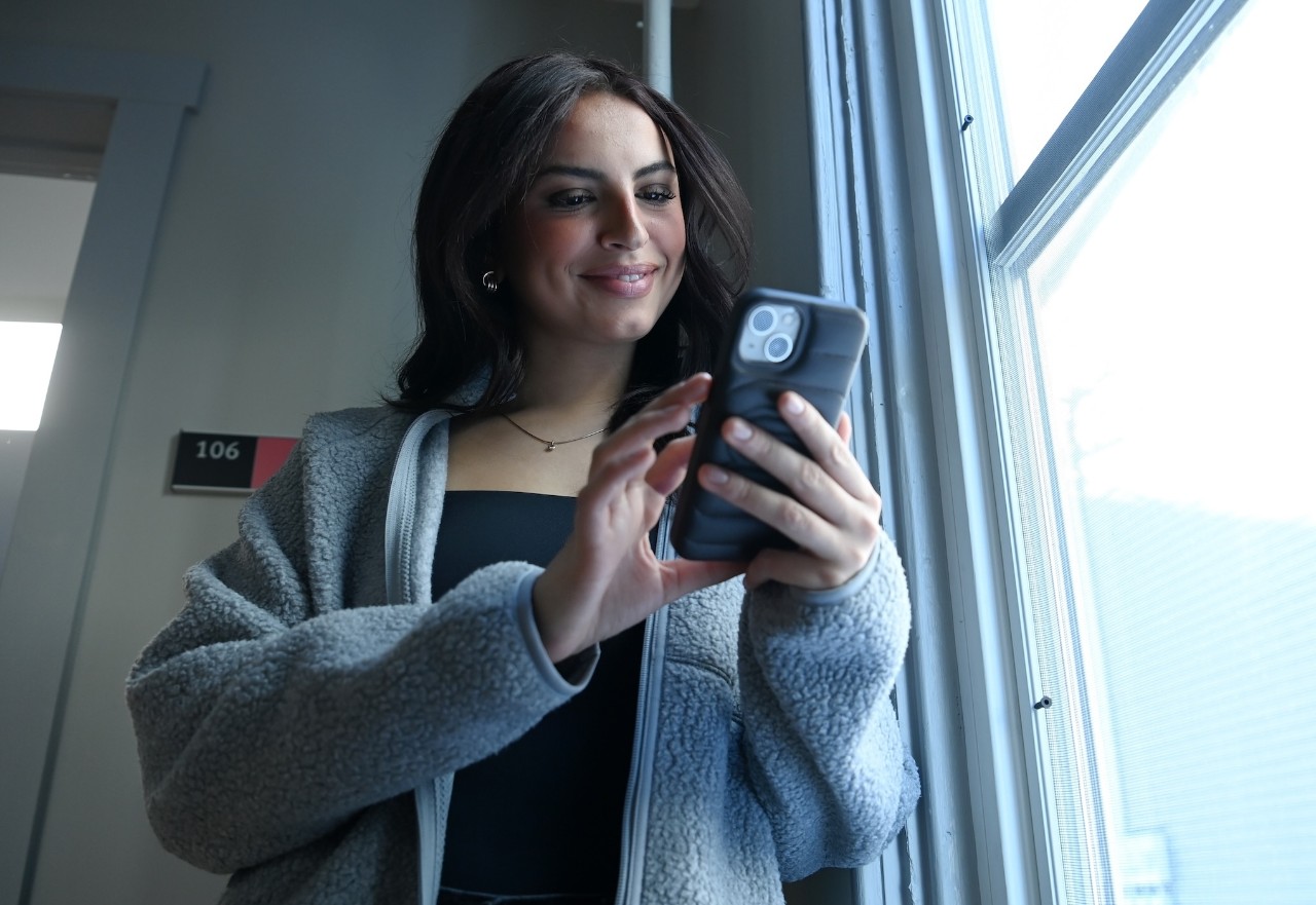 A student with long dark hair, wearing a black top and grey cardigan, stands in front of a window and looks at the mobile phone in her hands.